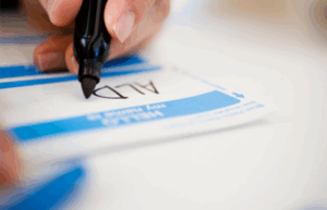 Close-up of a hand writing 'ALD' on a blue and white name tag with a black marker, emphasizing the theme of name recognition and memory training.