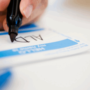 Close-up of a hand writing 'ALD' on a blue and white name tag with a black marker, emphasizing the theme of name recognition and memory training.